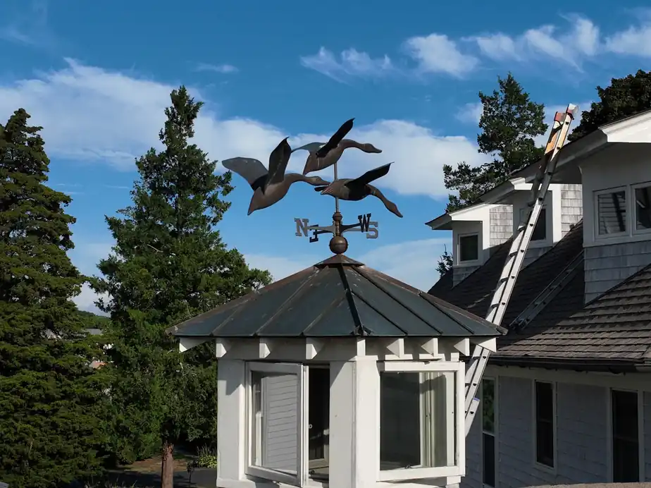 Painter on a ladder beside a white gazebo topped with a flying-geese weathervane, blue sky behind