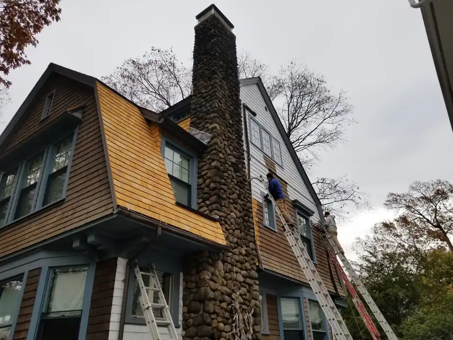 Shingled Dutch Colonial with a stone chimney and stained cedar siding, ladders up against the gable